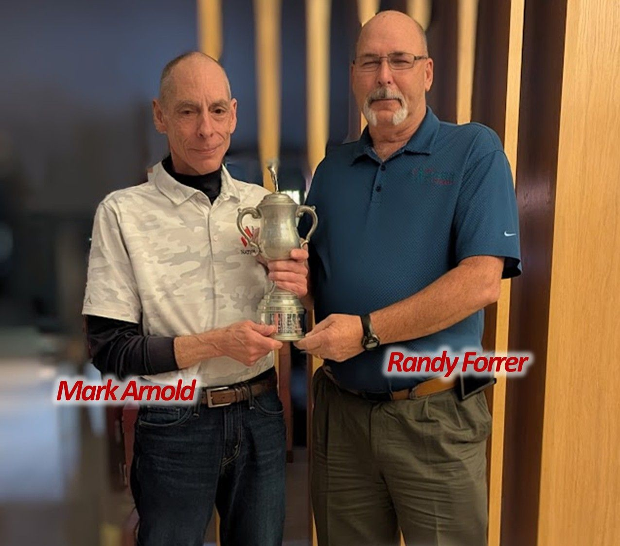Two men holding a trophy. Mark Arnold and Randy Forrer stand inside with a wooden wall behind them.