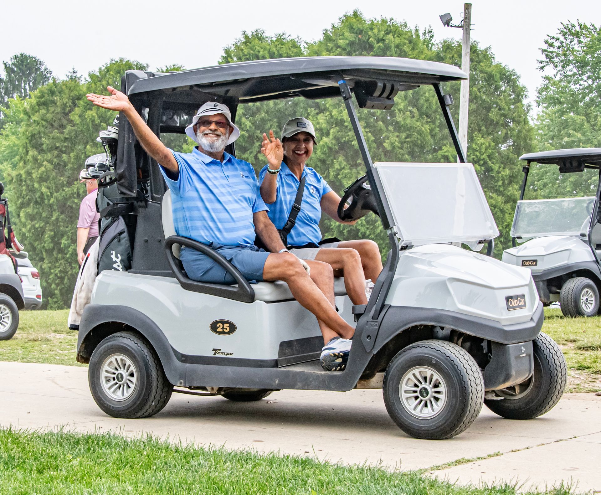 Two people are wavingsitting in a golf cart on a golf course.
