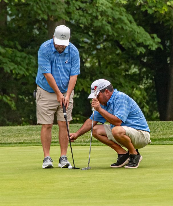 Two golfers on a green: one about to putt, the other squats examining the line.