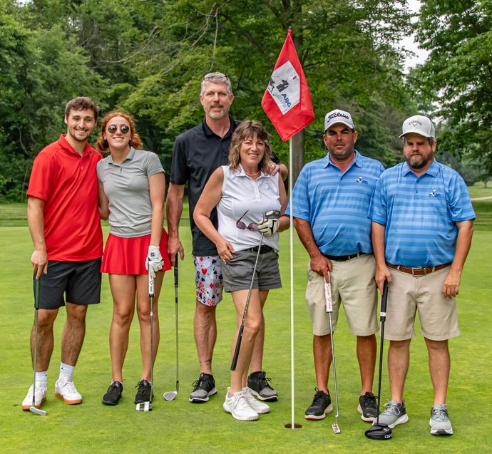 Group of golfers on a course. Six people pose near a red flag. Mostly sunny, green grass.