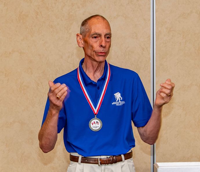 Mark Arnold in blue shirt with medal, giving speech; beige wall in the background.