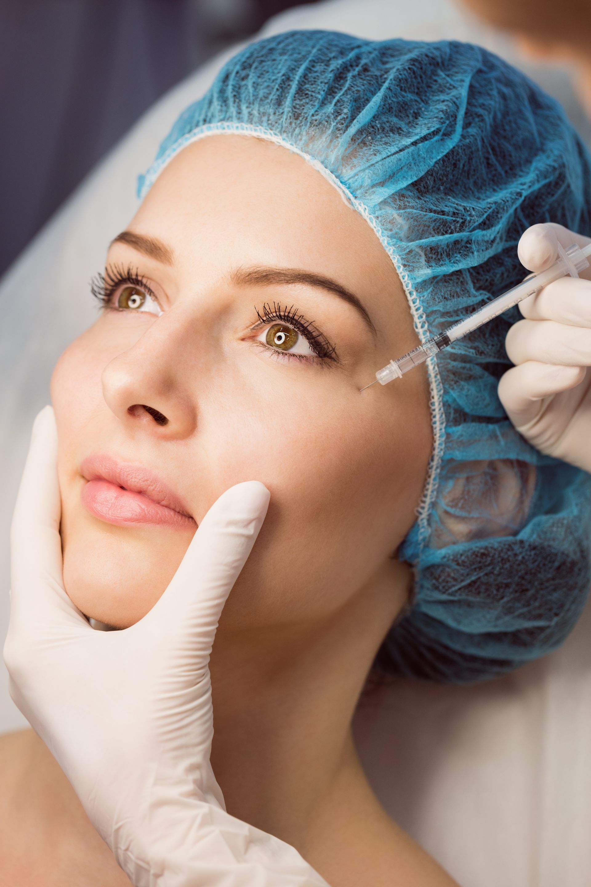 Person receiving cosmetic injections under the eye; hands in gloves, blue cap.