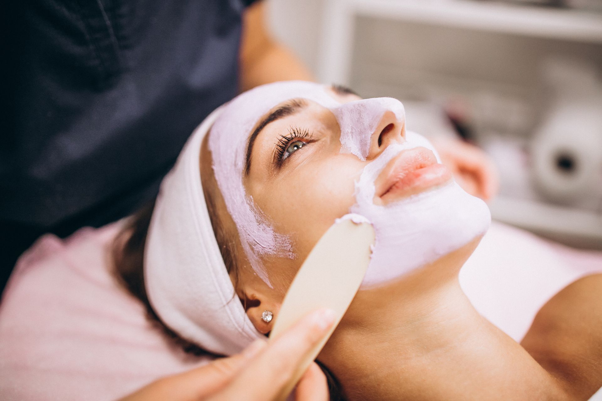 Woman receiving facial treatment, cream applied to face with spatula.