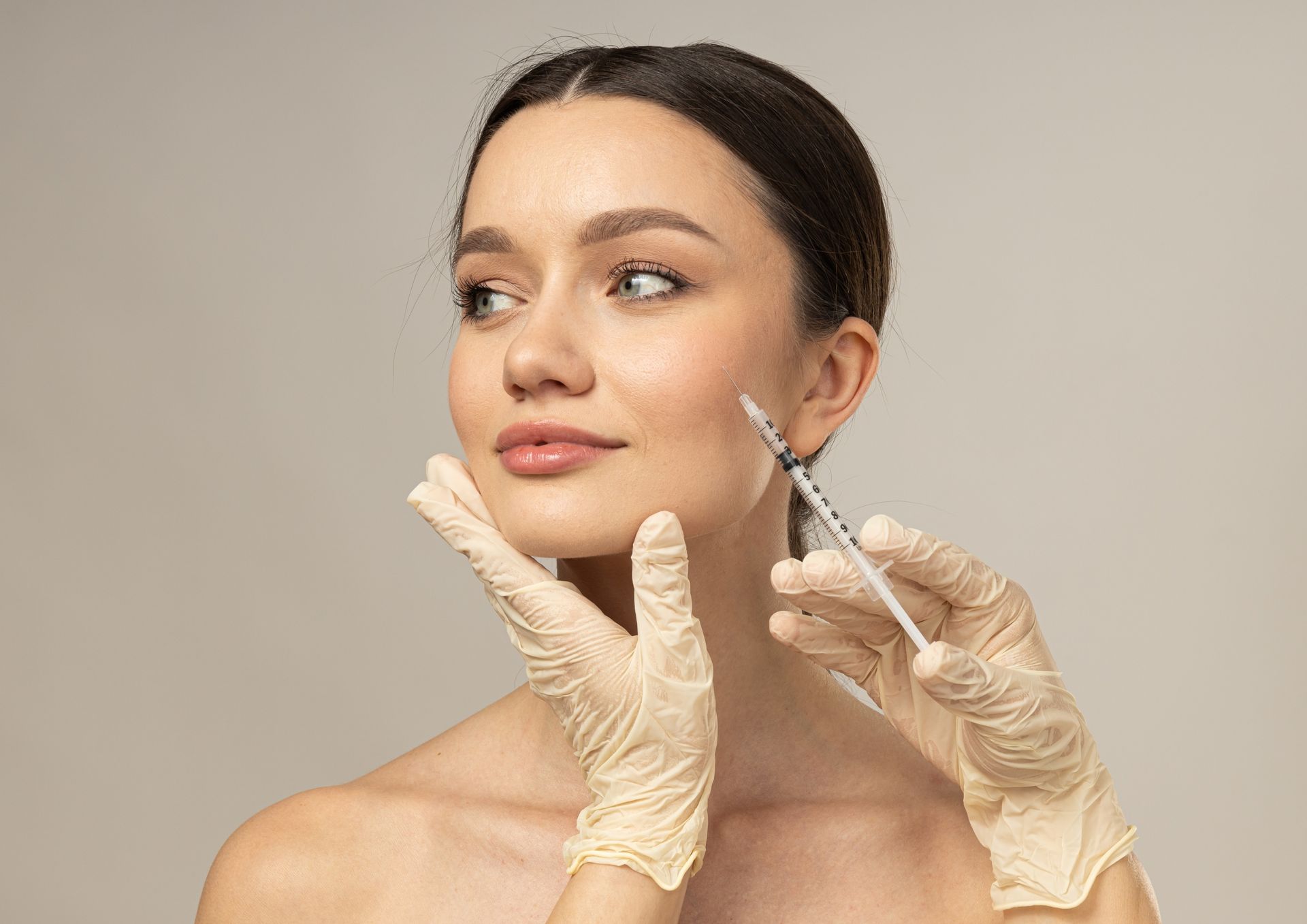 Person receiving a facial injection. A gloved hand holds a syringe near the cheek; light skin, neutral background.