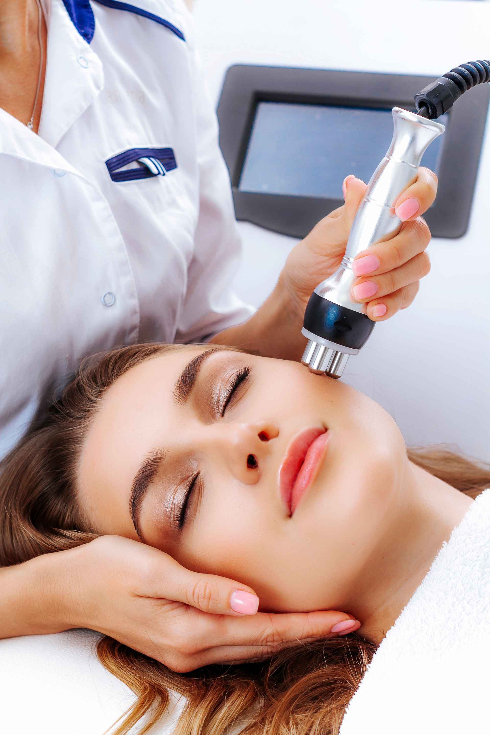 Woman receiving facial treatment with device; clinic setting.