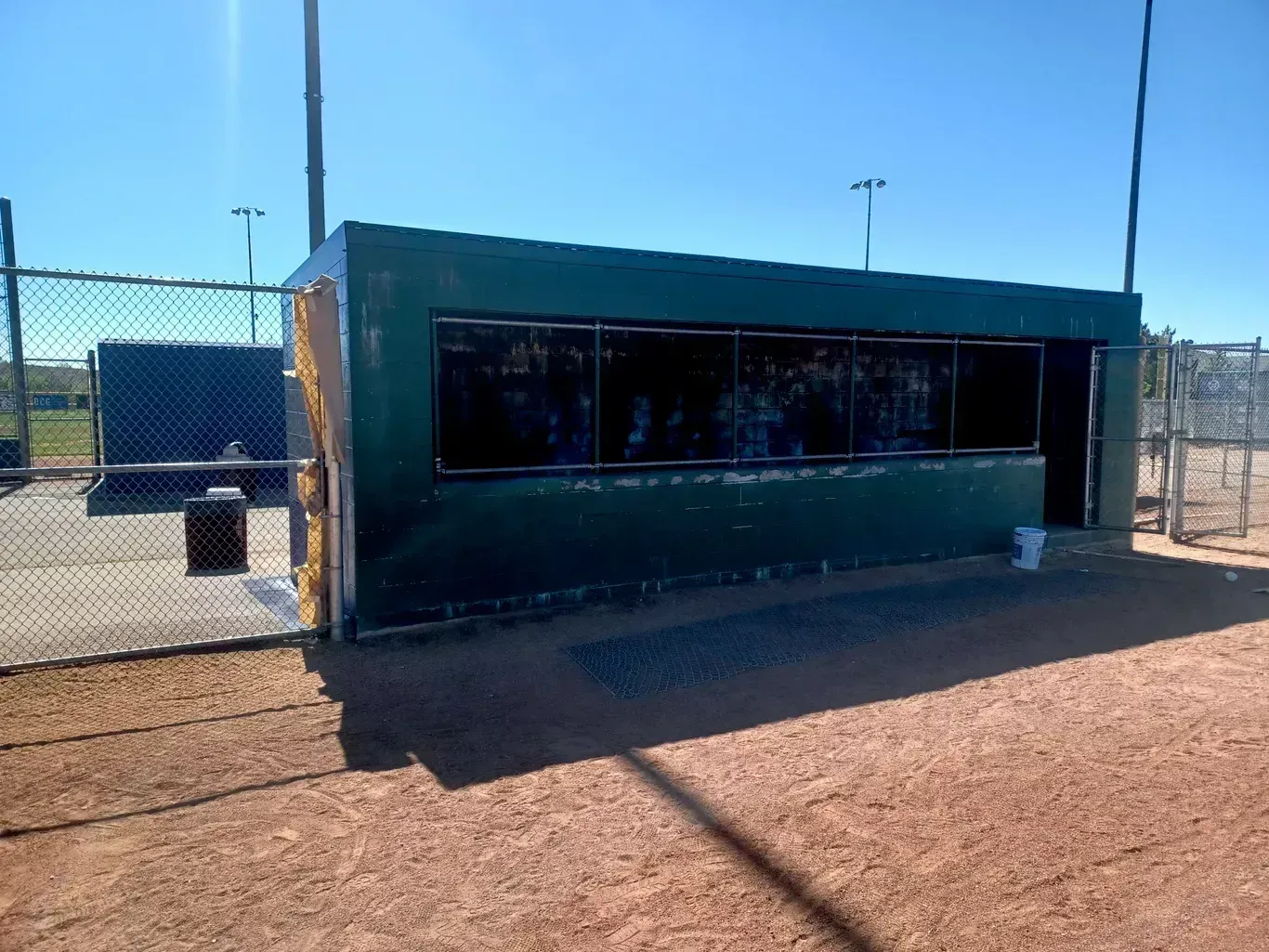 Small green concession stand with dark windows on a sunny baseball field, behind a chain-link fence.