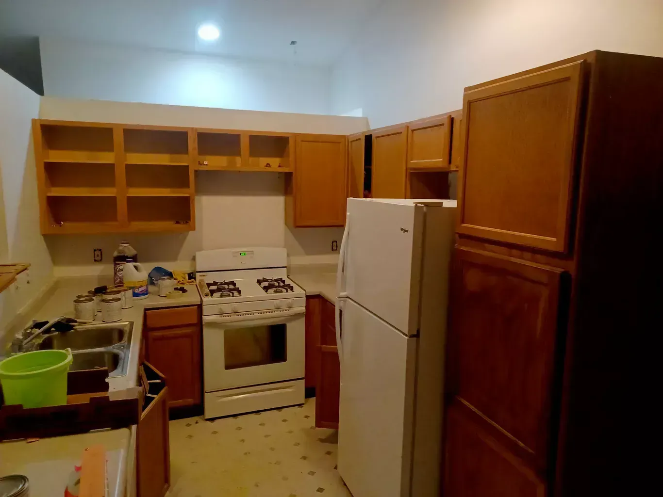 Small kitchen with wooden cabinets, white fridge, stove, and sink under a bright ceiling light.