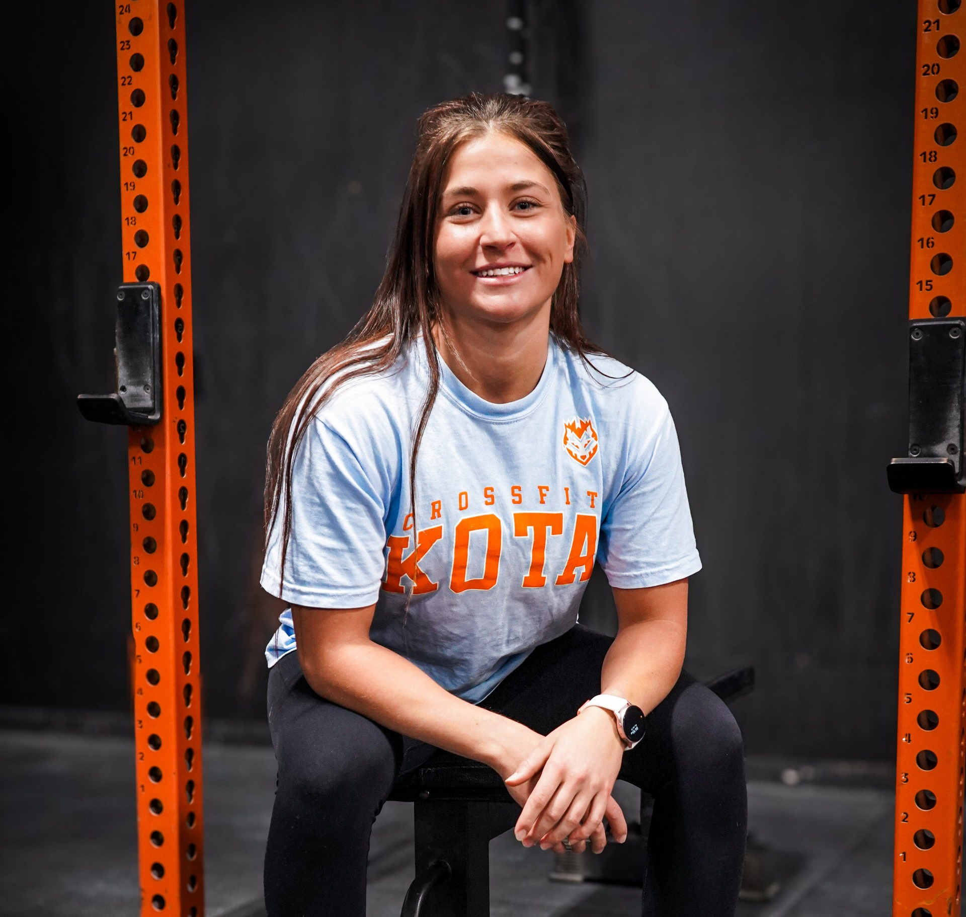 Woman in gym attire sits smiling, dumbbells in front. Orange gym equipment visible.