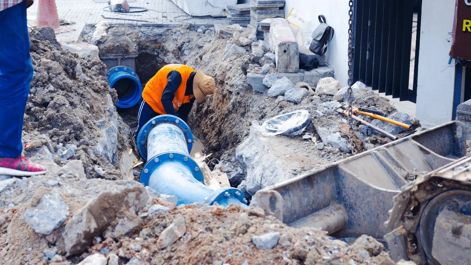 Workers in orange vests repairing a large blue pipe in an open trench, amidst debris and an excavator.