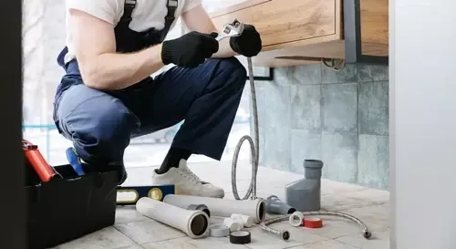 Plumber in overalls, kneeling under a sink, working with a wrench. Tools and pipes are nearby.