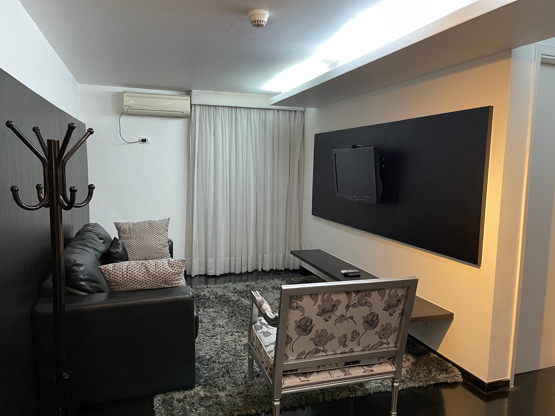 Uma sala de estar contemporânea com um sofá preto, uma poltrona estampada, uma TV fixada na parede e um cabideiro.