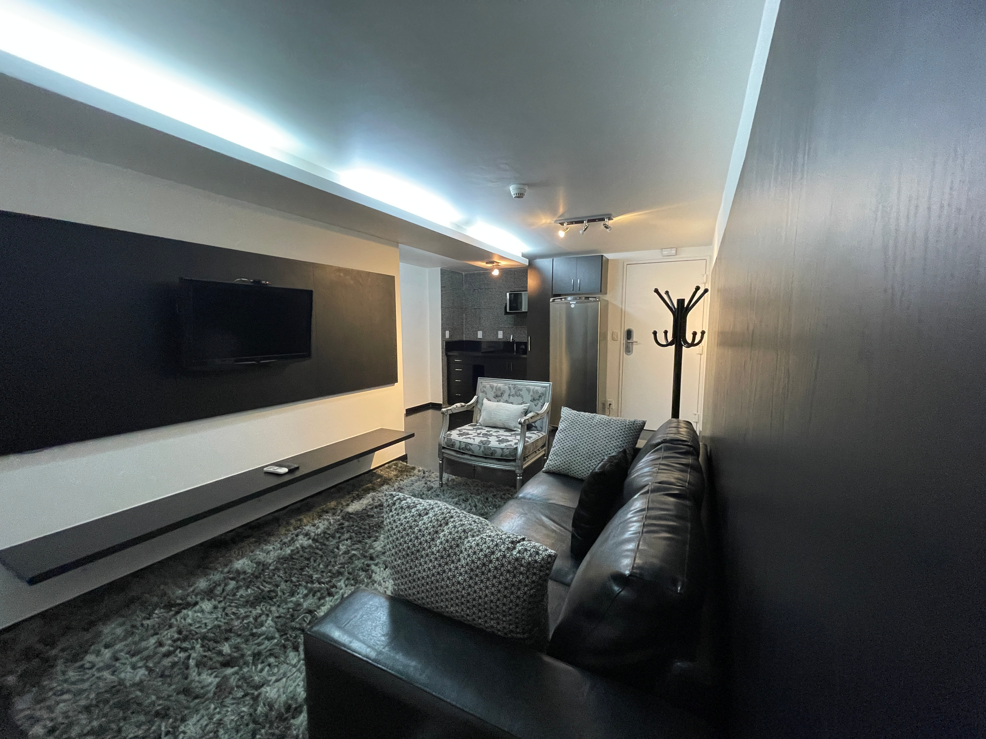 Uma sala de estar moderna com um sofá de couro escuro, poltrona, tapete felpudo cinza e uma TV montada na parede sobre uma longa prateleira suspensa.