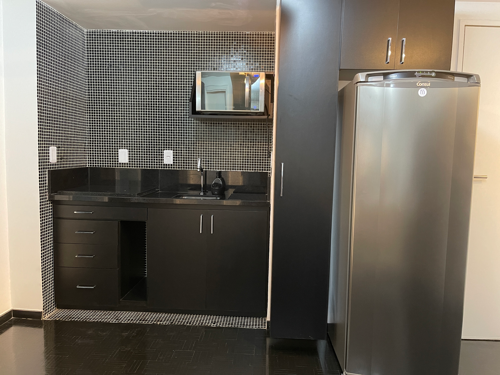 Pequena cozinha americana com armários escuros, geladeira de aço inoxidável e backsplash com padrão quadriculado em preto e branco.