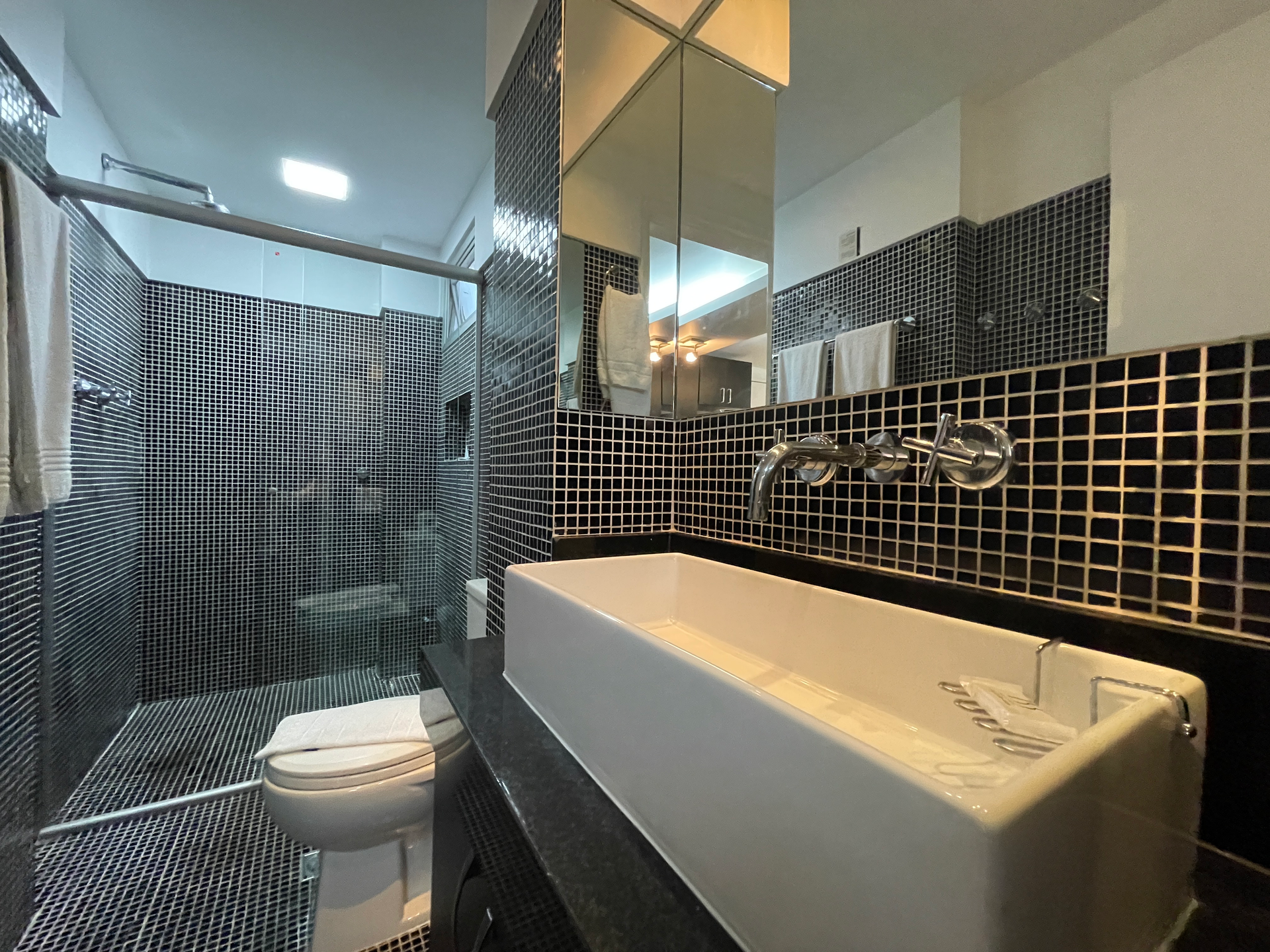 Banheiro moderno com pia retangular branca, parede revestida com azulejos pretos, bancada com espelho e box de chuveiro com estampa em preto e branco