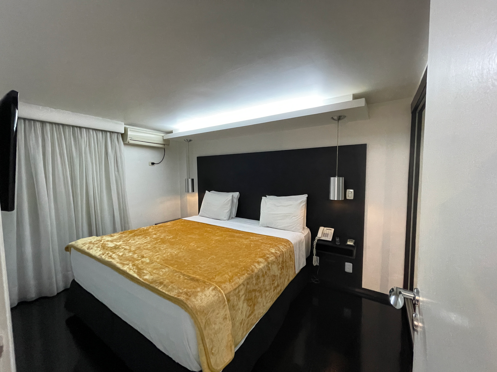 Um quarto de hotel com uma cama coberta de ouro, cabeceira preta, cortinas brancas e um ar-condicionado fixado na parede.