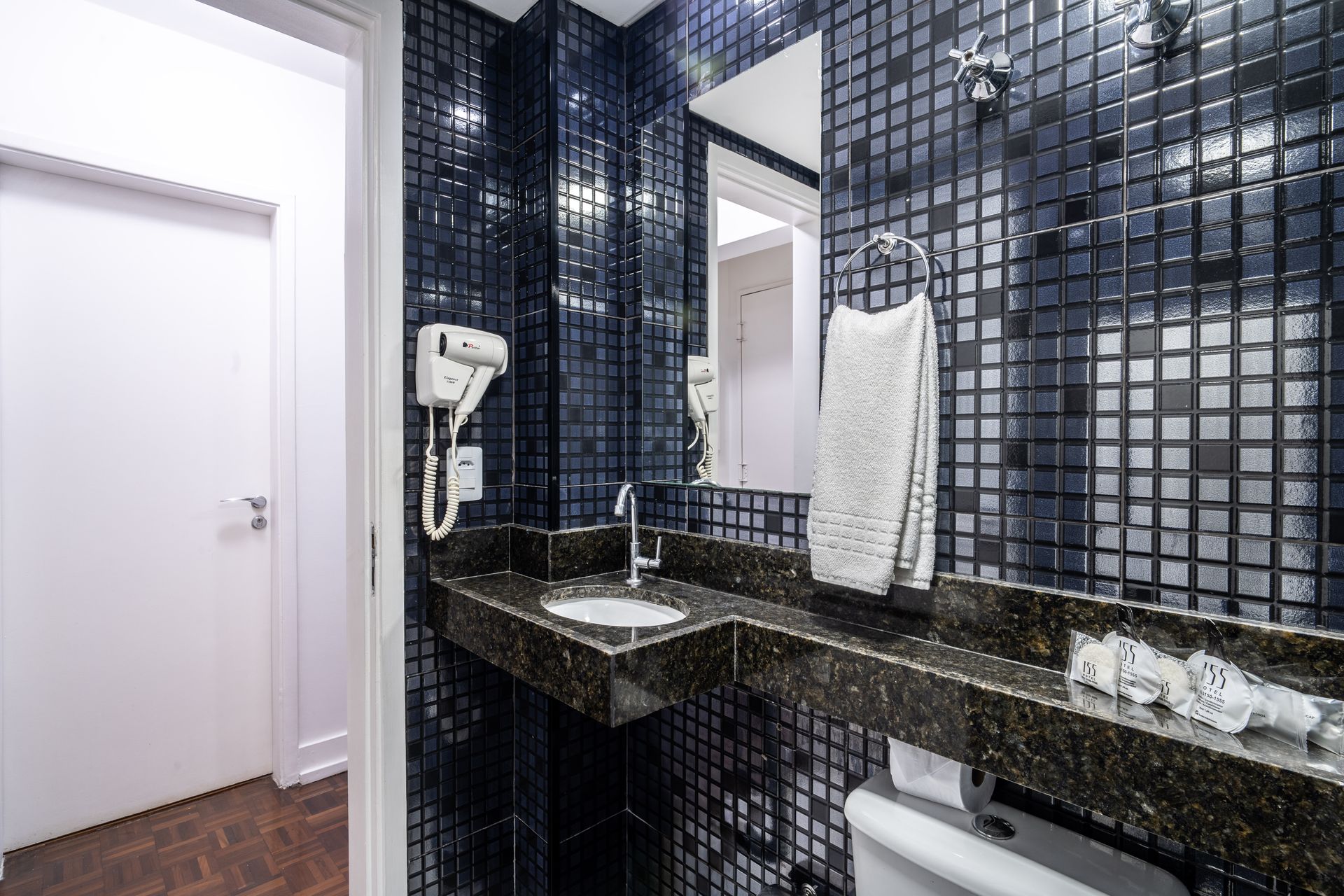 Banheiro com paredes revestidas com azulejos de mosaico azul escuro, bancada de pedra escura, pia branca, secador de cabelo fixado na parede e espelho.