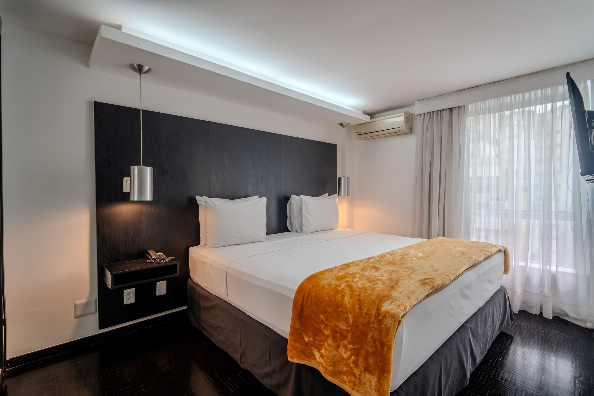 Um quarto com cama queen-size, cabeceira de madeira escura, luminária pendente e cortinas translúcidas na janela.