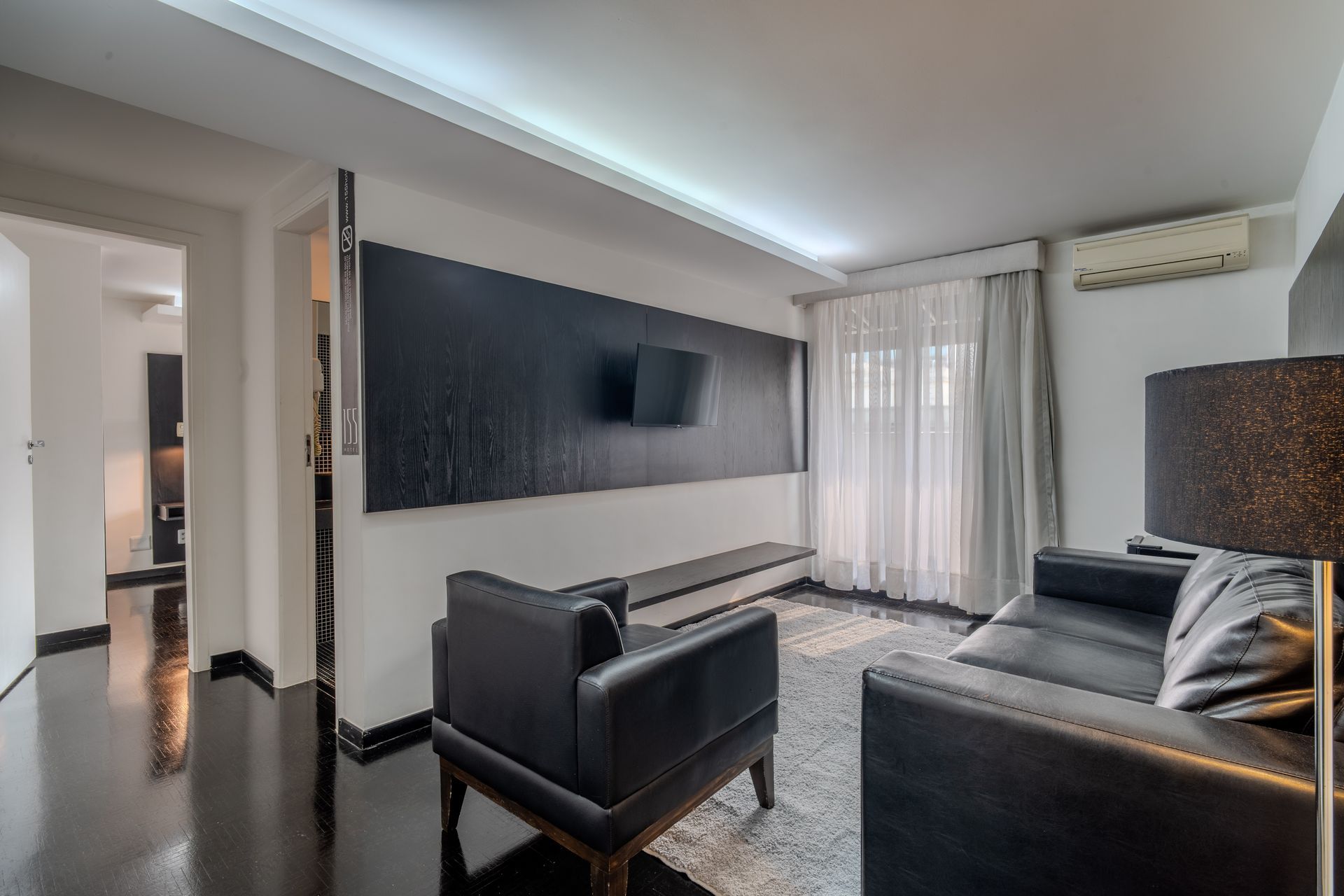 Sala de estar moderna com poltrona e sofá de couro preto, uma parede de destaque escura com TV e um tapete branco.