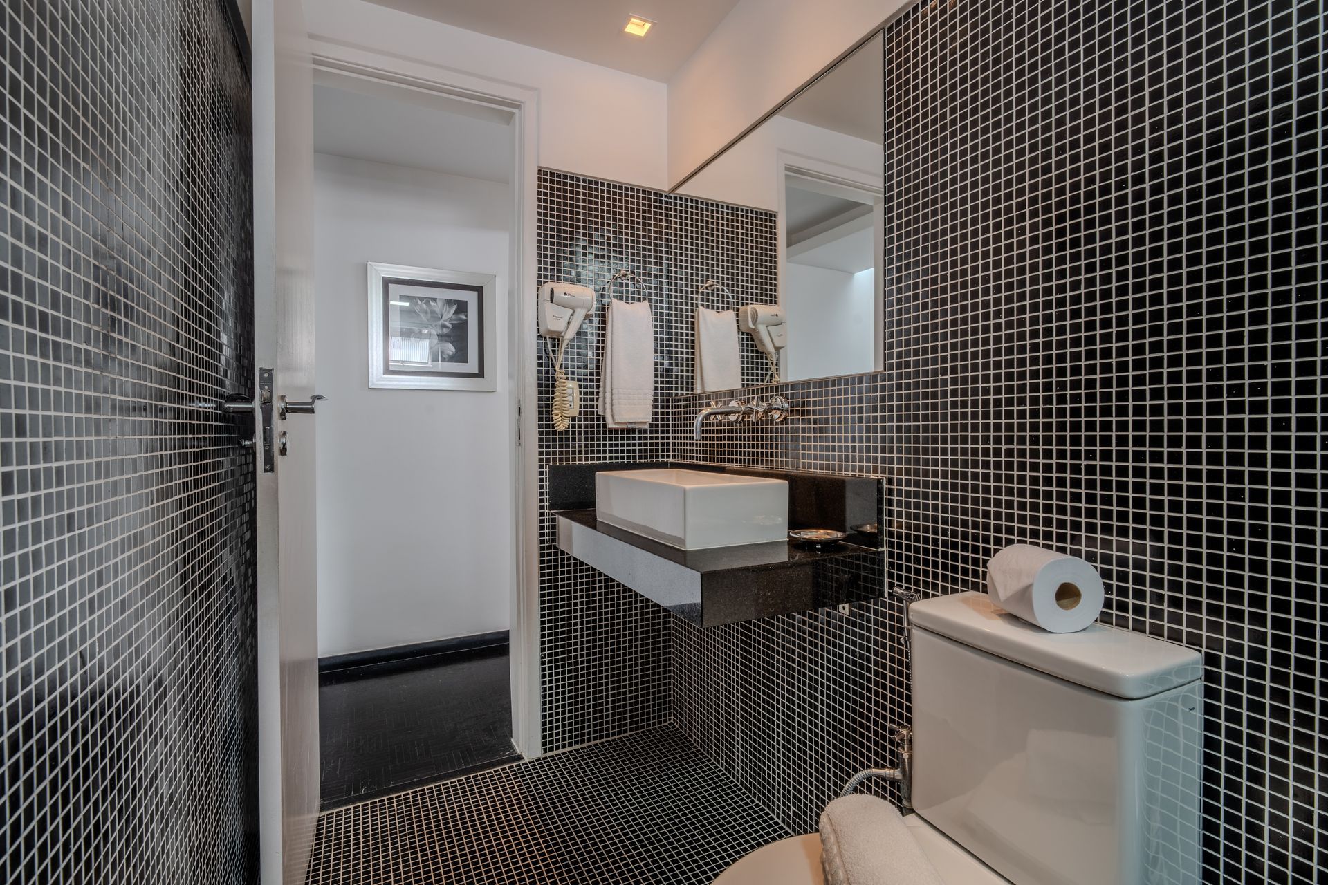 Um pequeno banheiro com paredes revestidas de azulejos em mosaico preto e branco, uma pia retangular sobre uma bancada escura e um vaso sanitário branco.