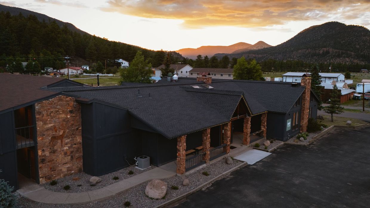 Stone lodge with dark roof at sunset, set among trees and mountains near a rural road.