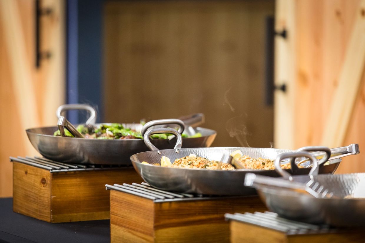 Buffet-style food in metal pans on wooden stands, with a blurred wooden background