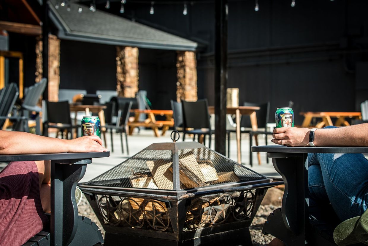 Two people sit beside a outdoor fire pit on a patio with tables and chairs nearby.