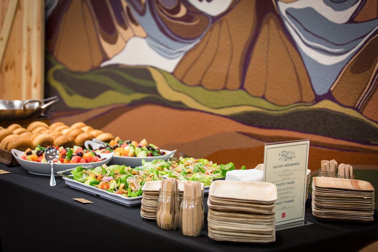 Buffet table with sandwiches, salads, fruit trays, napkins, and stacked plates in front of a mural backdrop