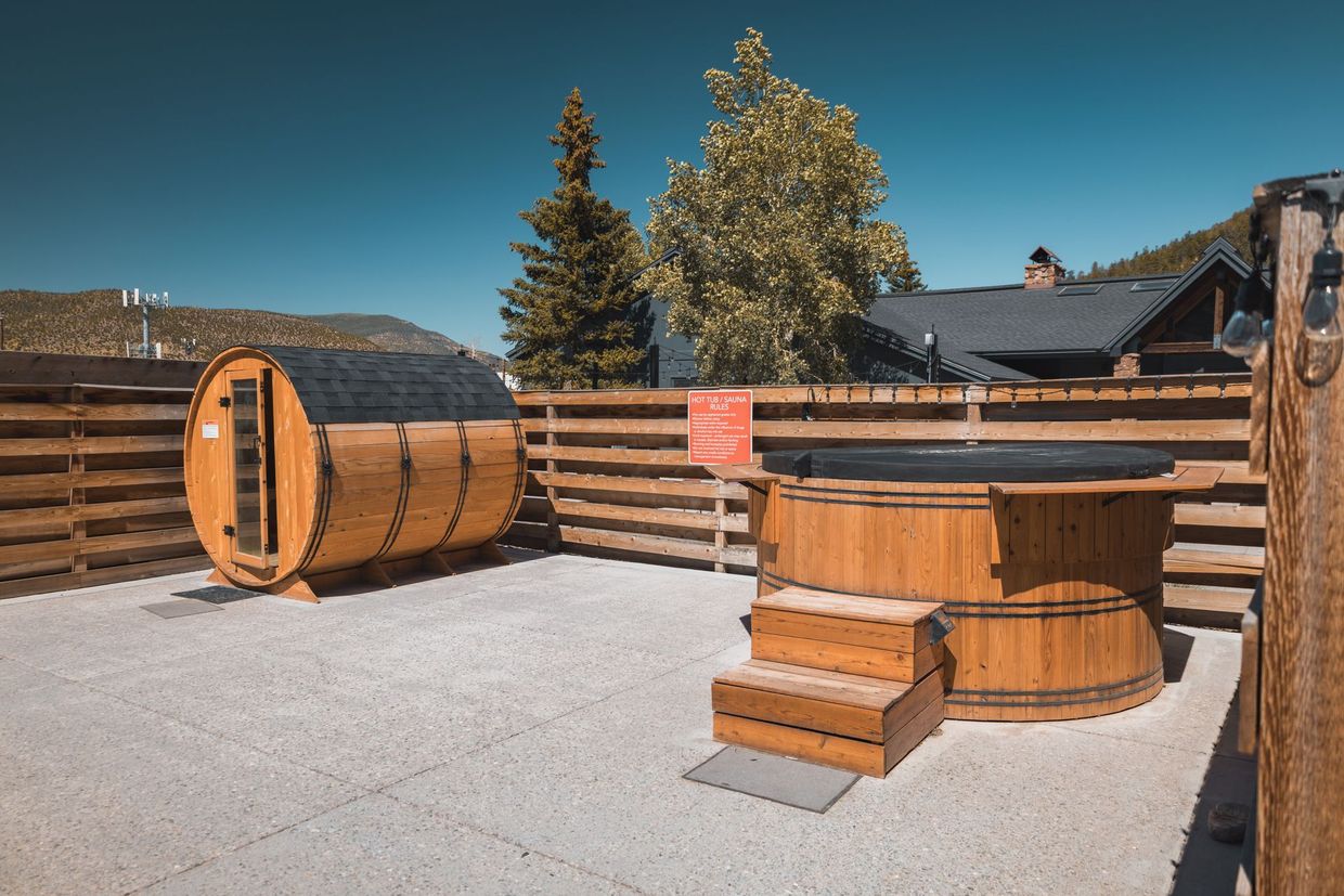 Outdoor wooden hot tub and barrel sauna in a fenced gravel yard under a clear blue sky