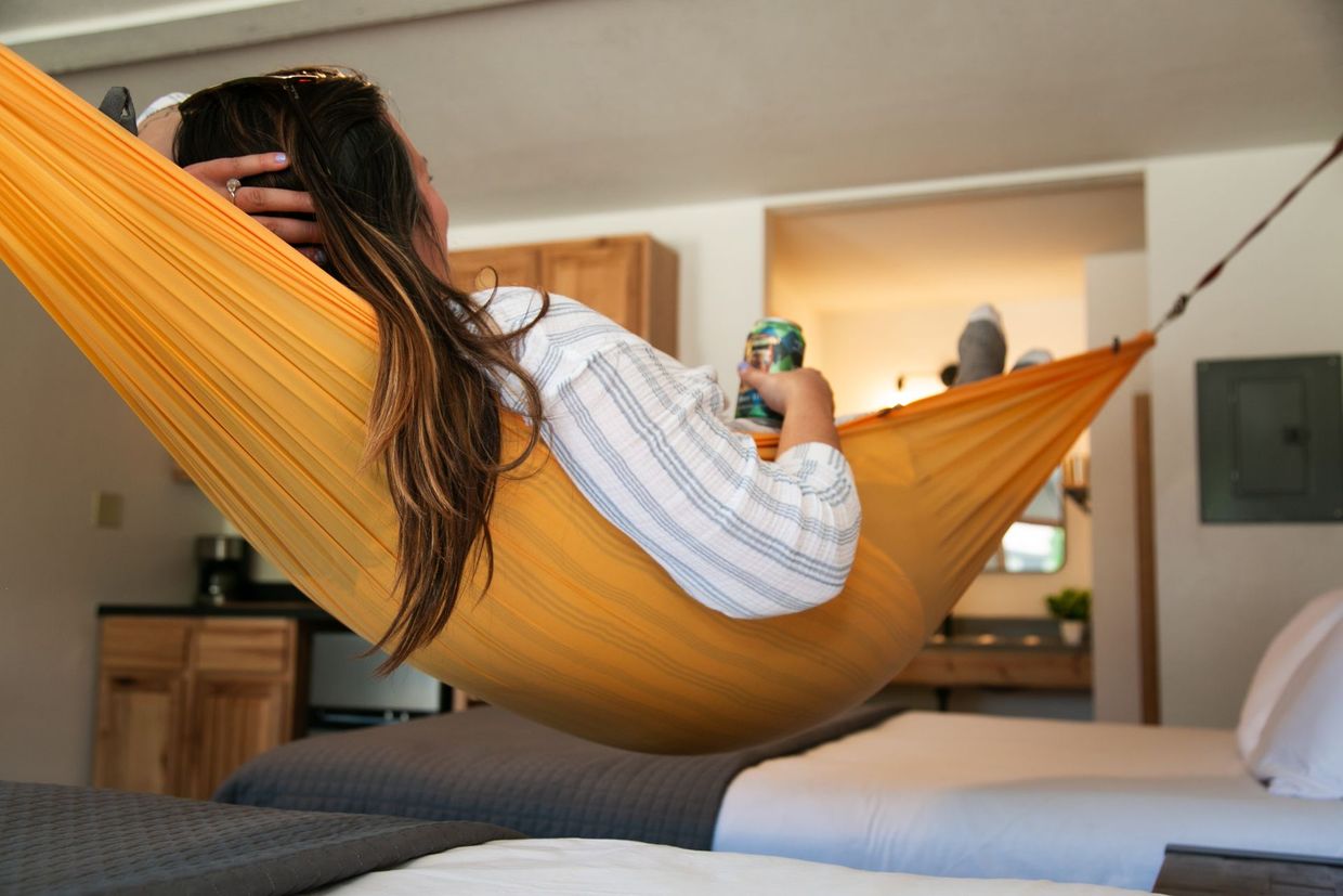 Person relaxing in an orange hammock in a bright bedroom, holding a drink.