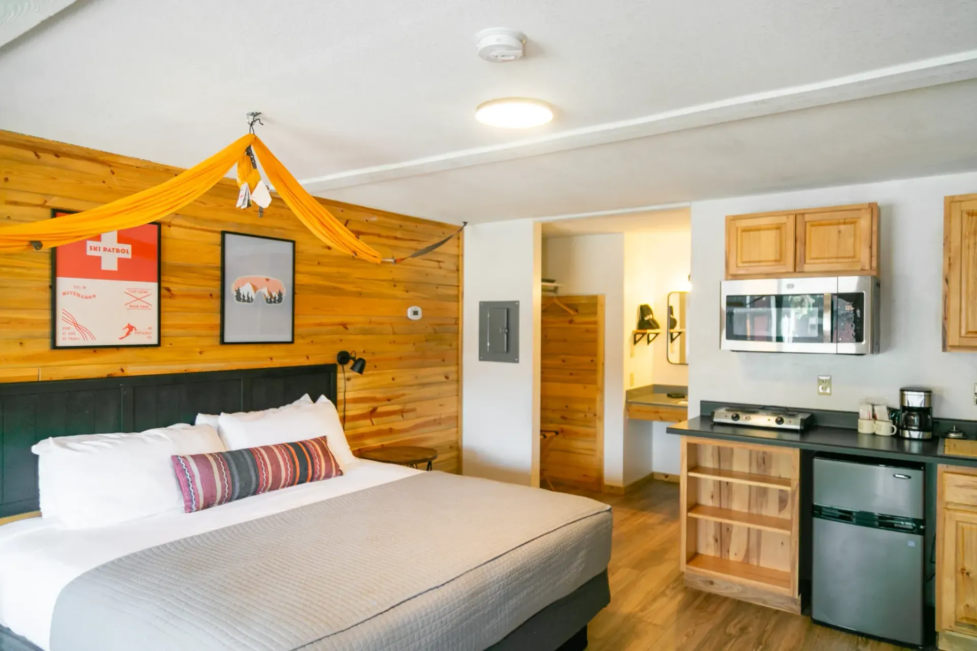 A hotel room with a queen bed, wooden accent wall, yellow decor, and a small kitchenette with a microwave and fridge.