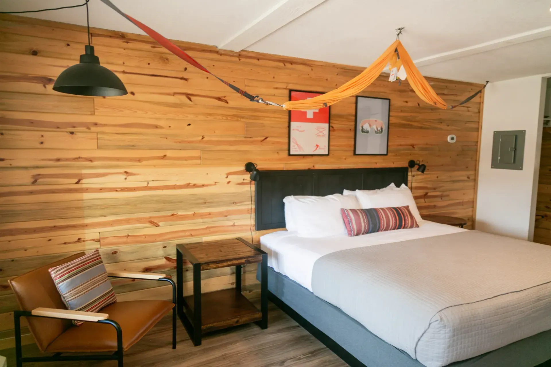 A bedroom with a bed, a wooden wall with two framed prints, a small side table, and a brown leather armchair.