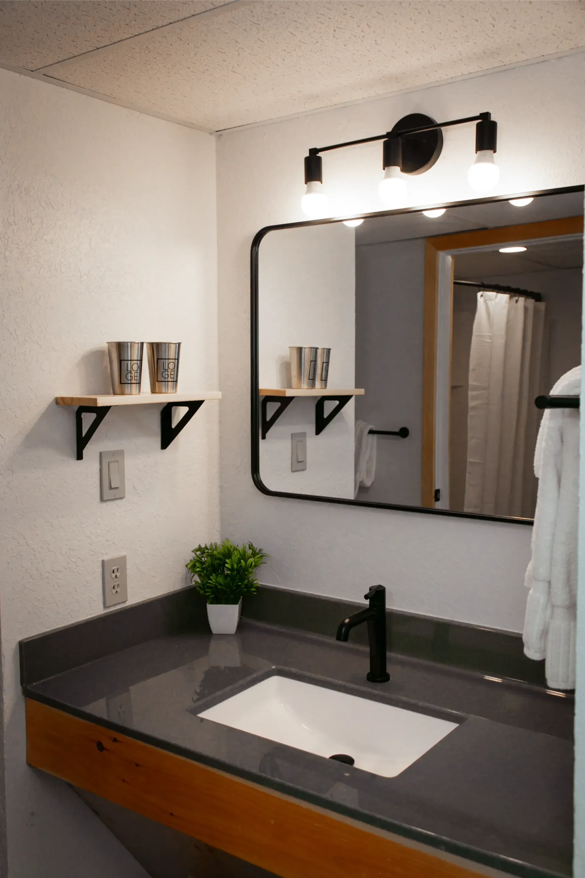 A modern bathroom vanity with a dark gray countertop, black faucet, white sink, and a floating shelf with silver cups.