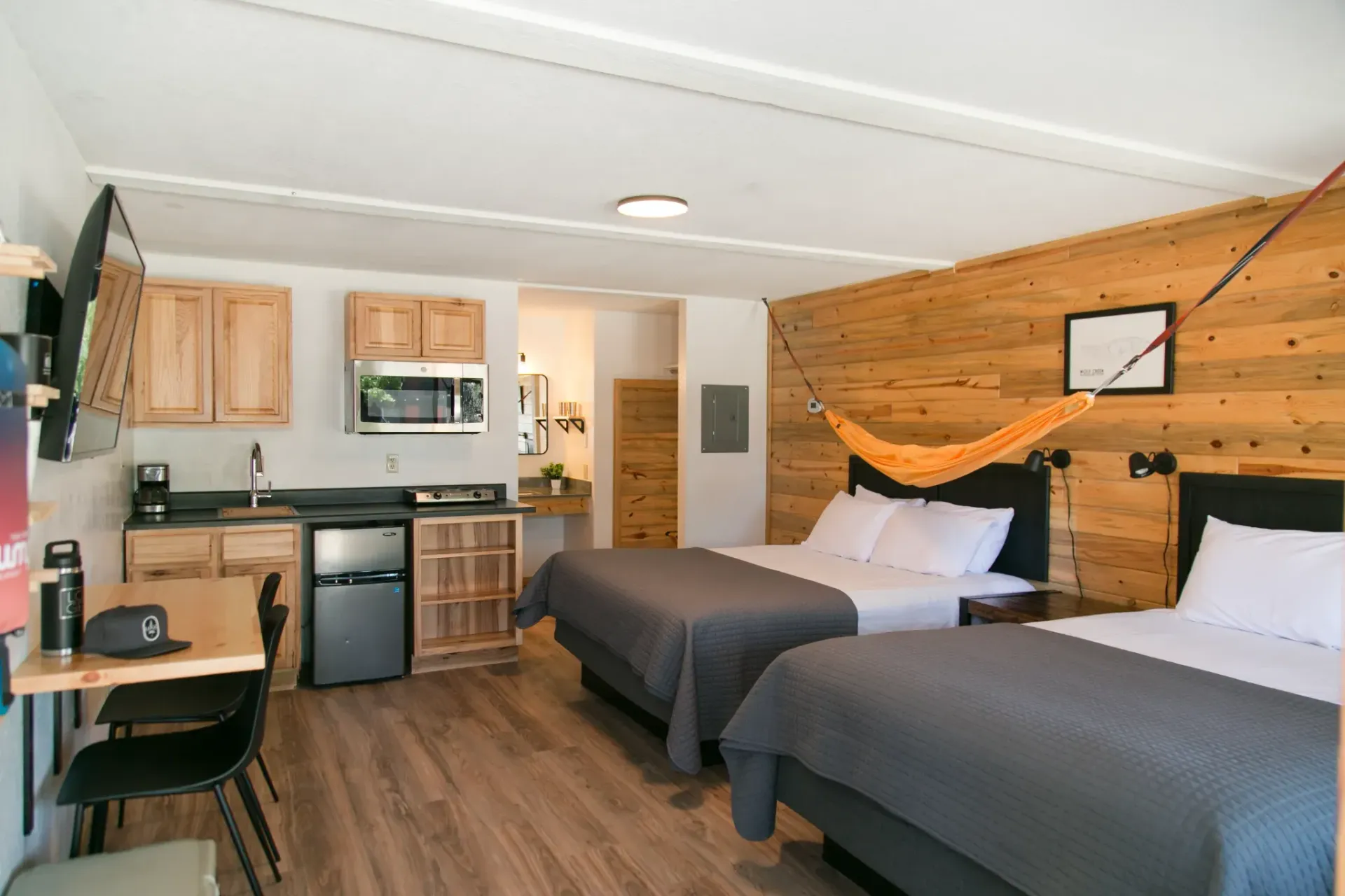 A hotel room with two beds, a small kitchen area with cabinets and appliances, and a hammock hung above the beds.