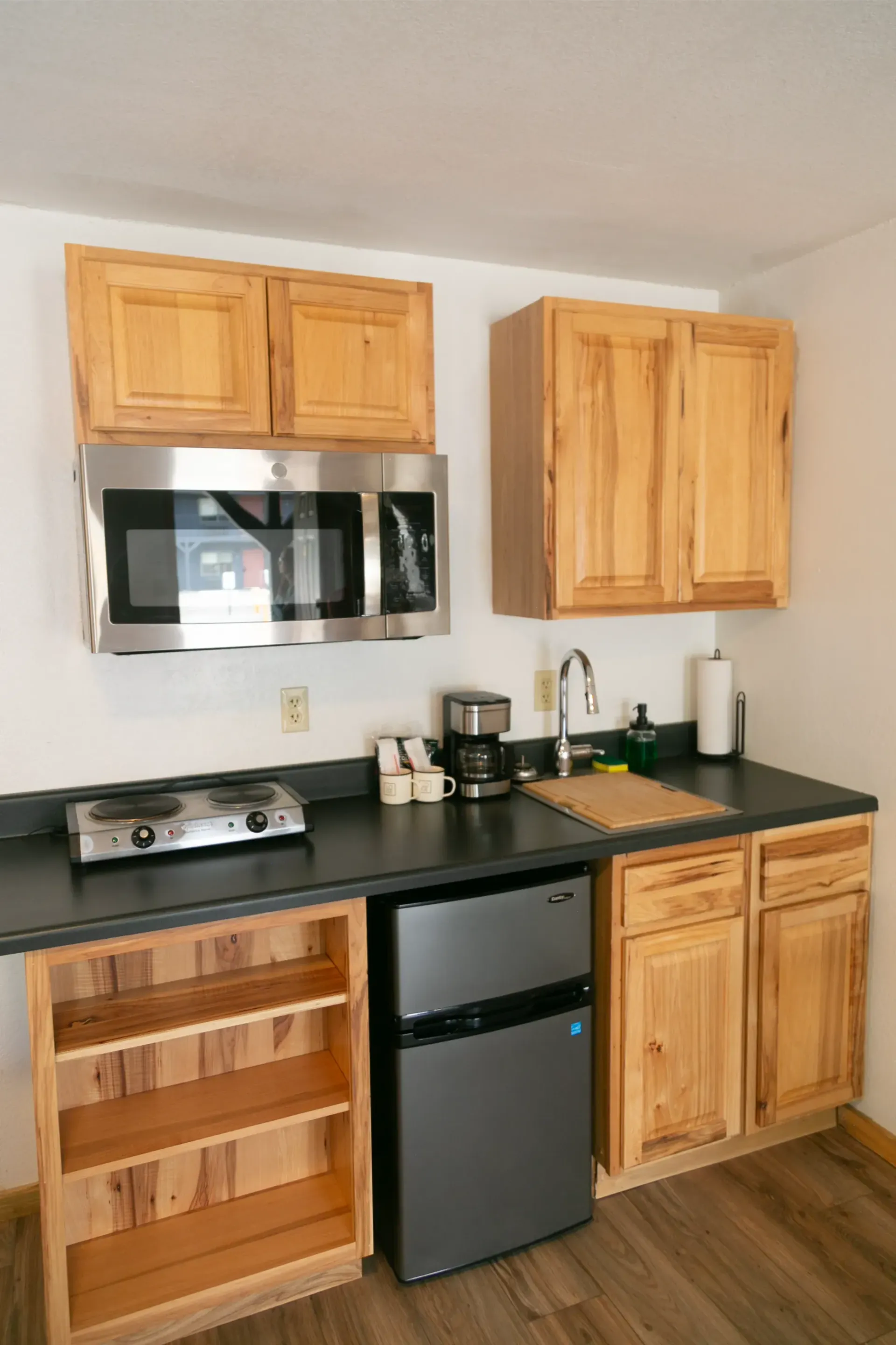 A compact kitchen with wooden cabinets, a small refrigerator, a microwave, and a stovetop on a dark countertop.