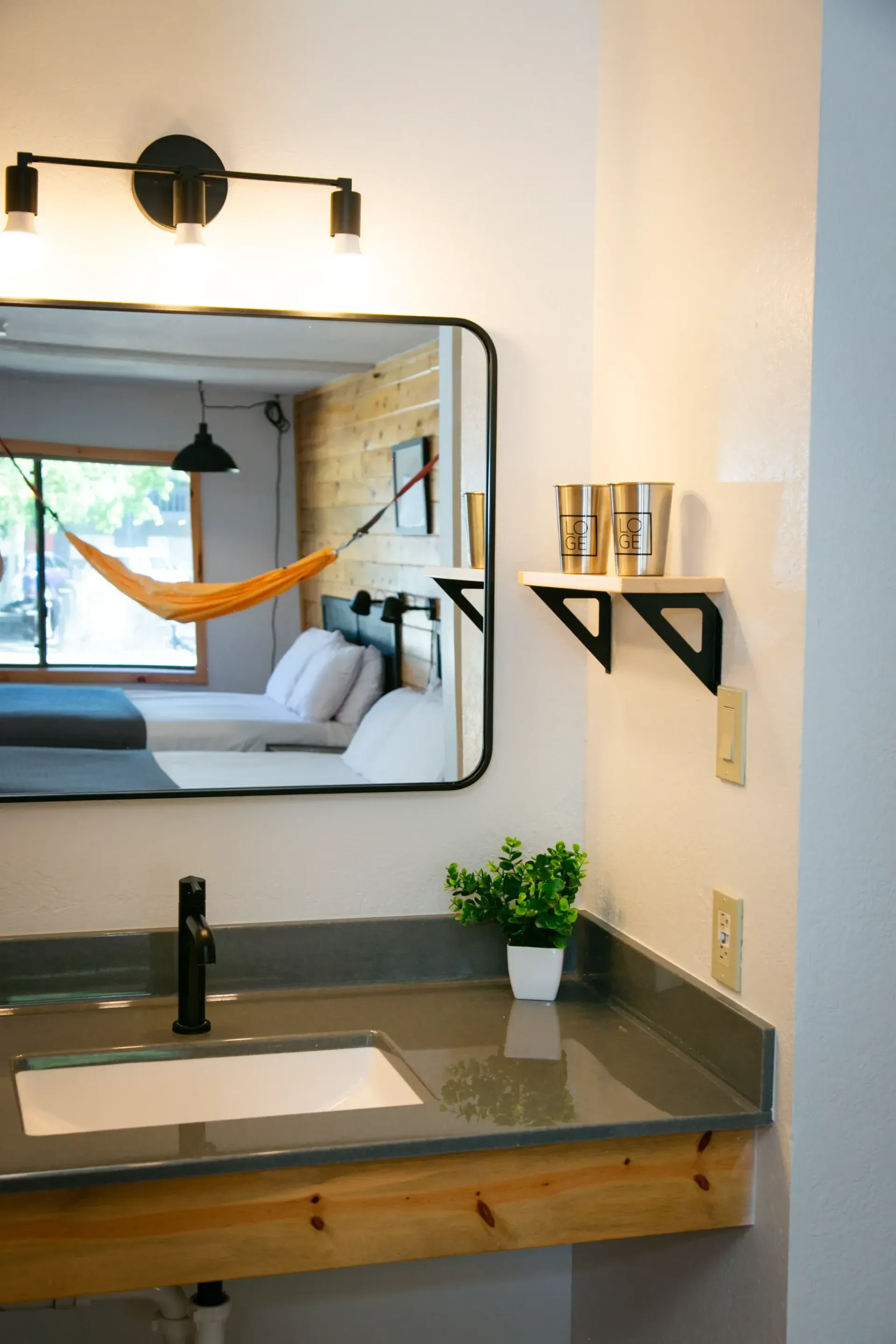A bathroom sink with a dark countertop, white basin, and mirror reflecting a bedroom with a hammock and wood-paneled wall.