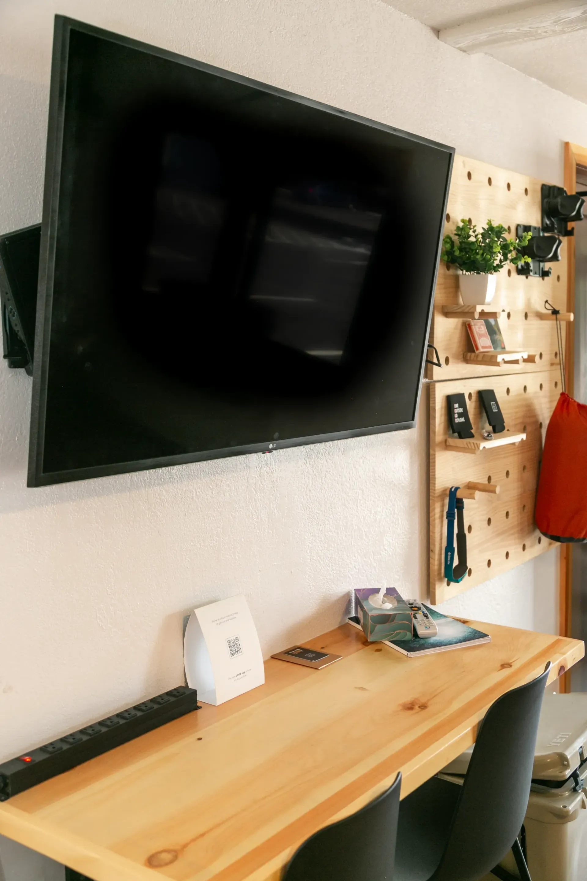 A wall-mounted TV hangs above a wooden desk and pegboard organizer in a bright, modern room.