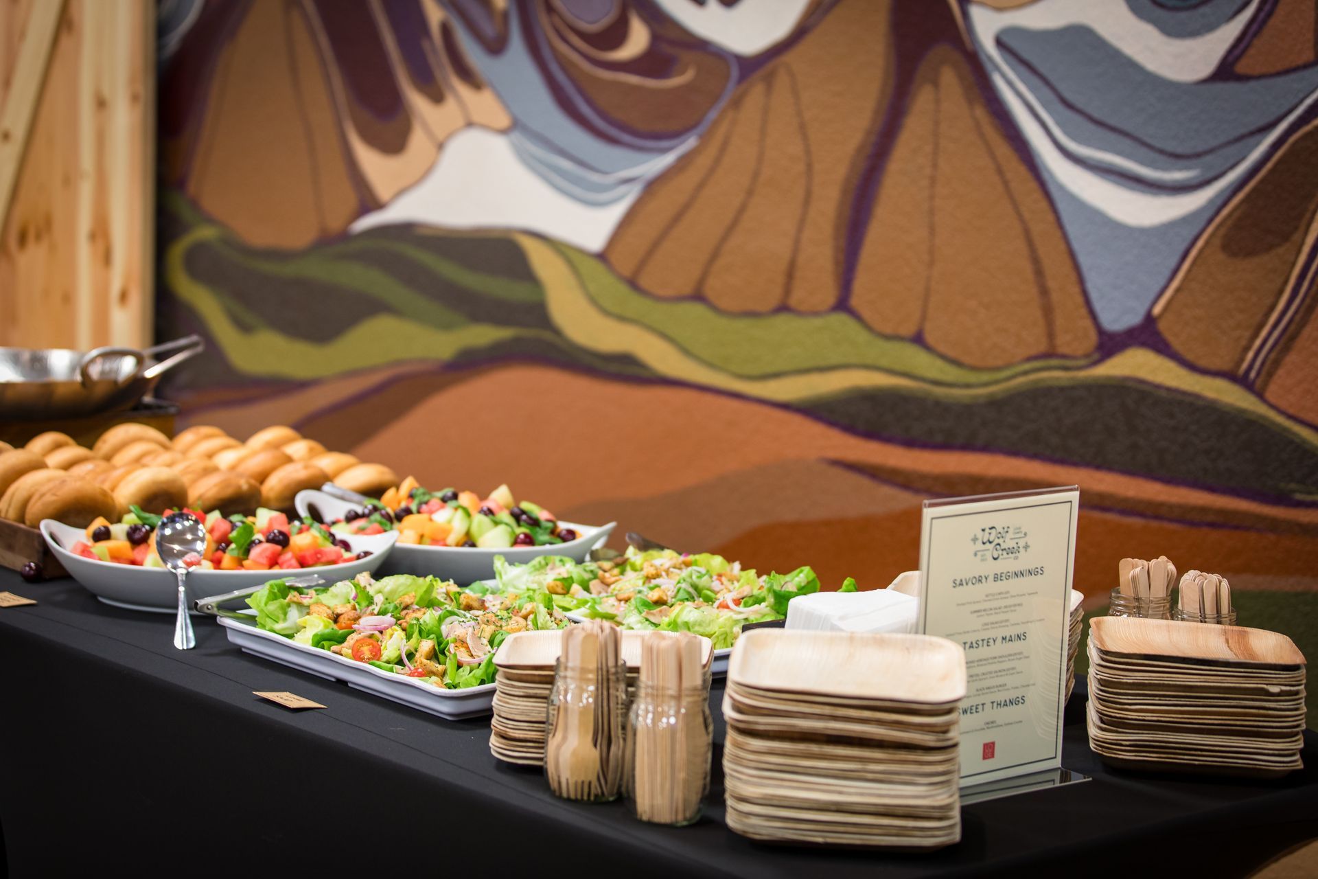 Buffet table with salads, bread rolls, pastries, and stacked plates in front of a mural backdrop