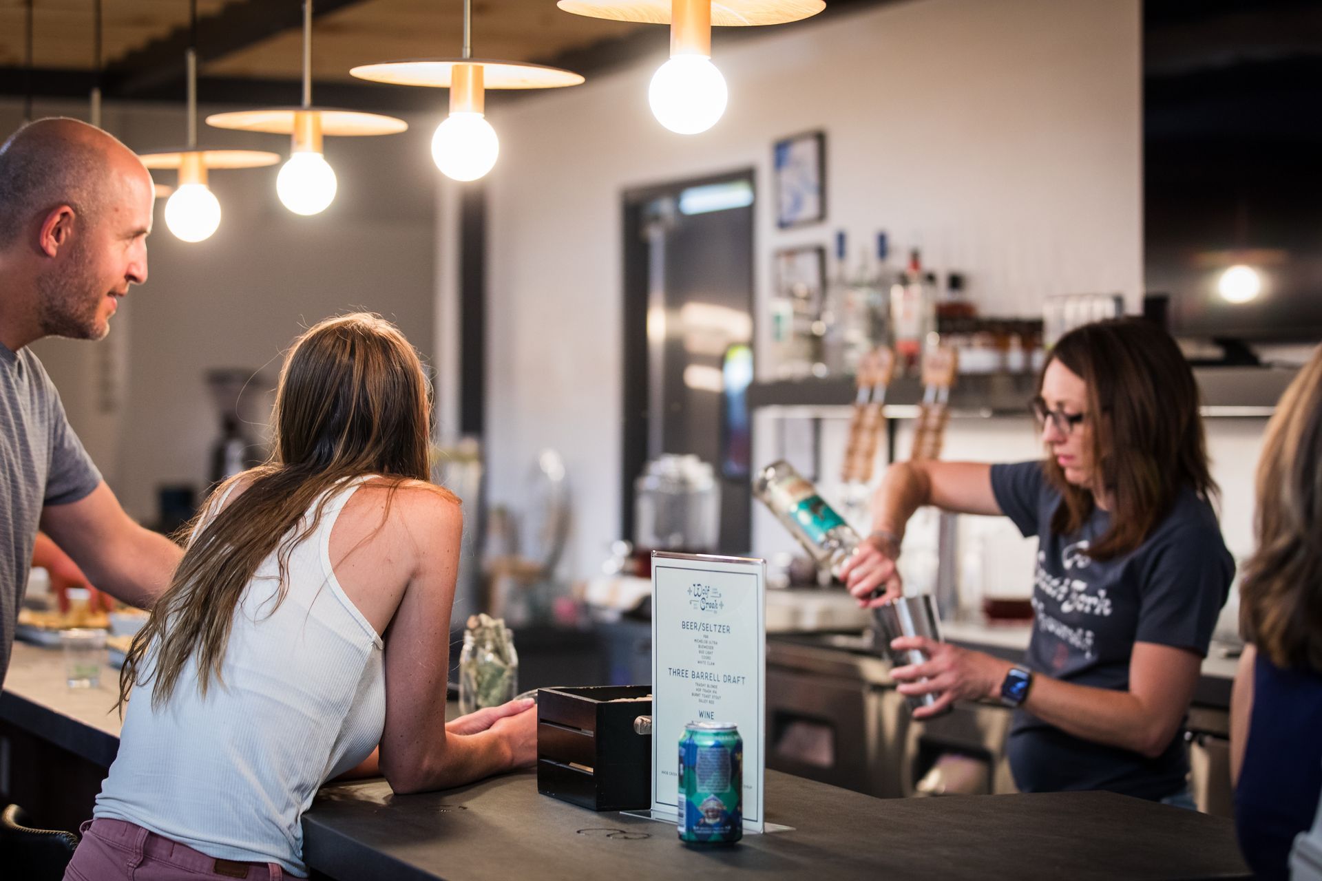 People chatting at a modern café counter under warm hanging lights, with drinks and a laptop on the counter