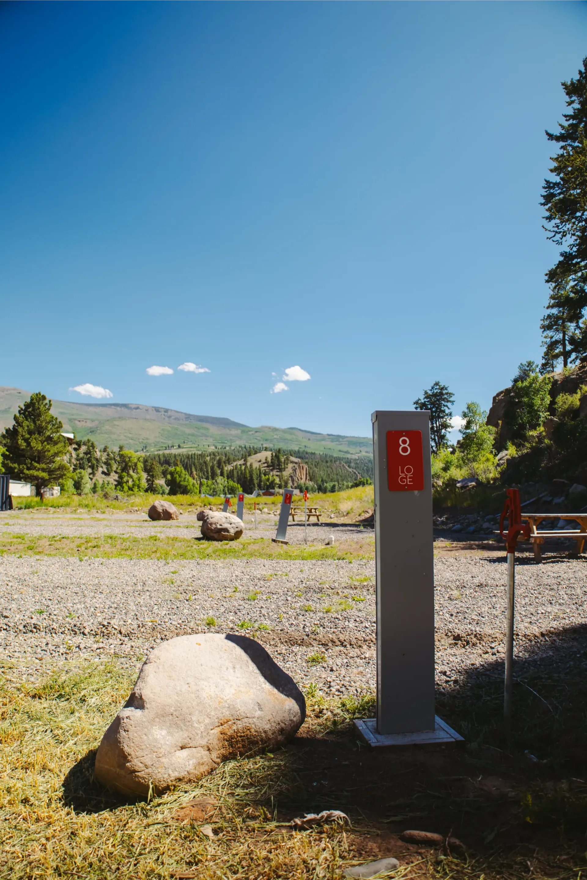 A red campsite number marker stands on a gravel campground with mountains, trees, and blue sky in the background.