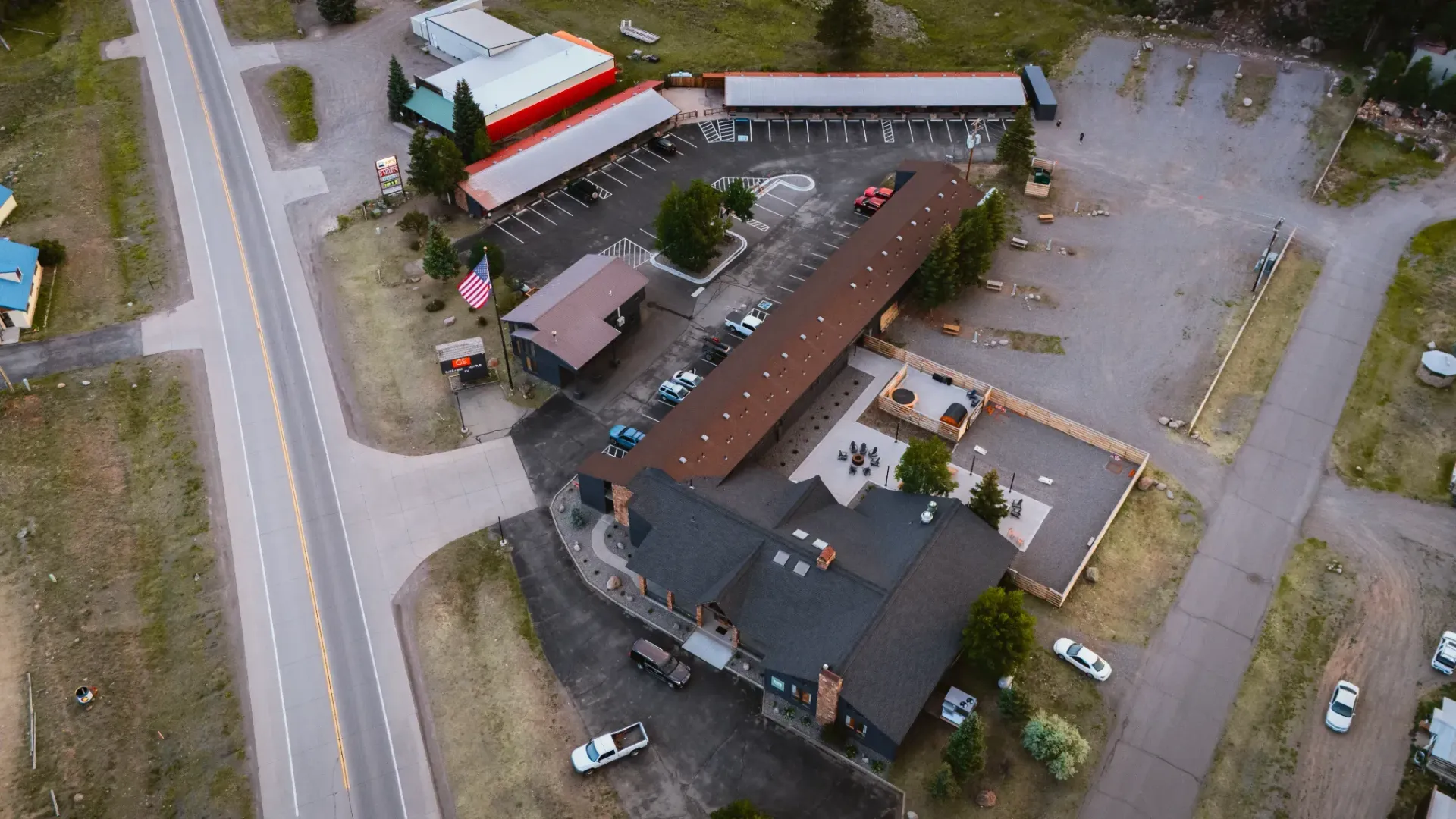 Aerial view of a lodge or motel complex with a long brown roof, parking areas, and a nearby road surrounded by trees.