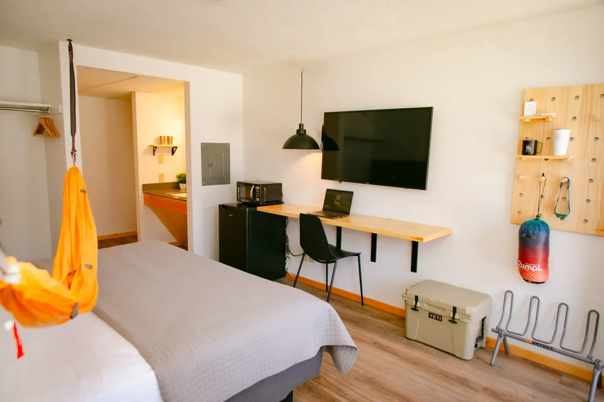 A hotel room with a bed, a desk area with a television, an orange hanging chair, a small refrigerator, and a gear wall.