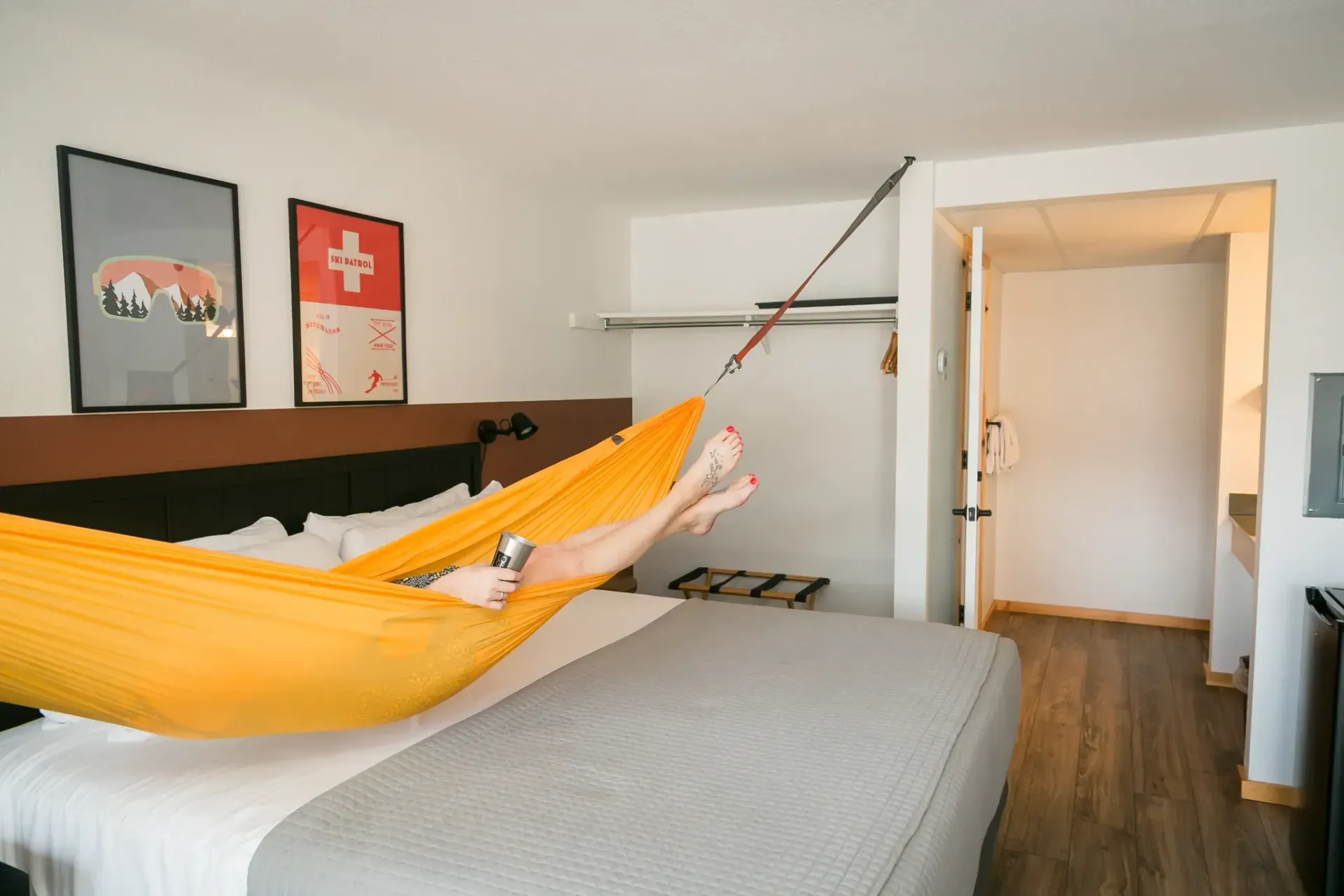 A person rests in a bright yellow hammock strung across a bed in a hotel room with two wall posters.