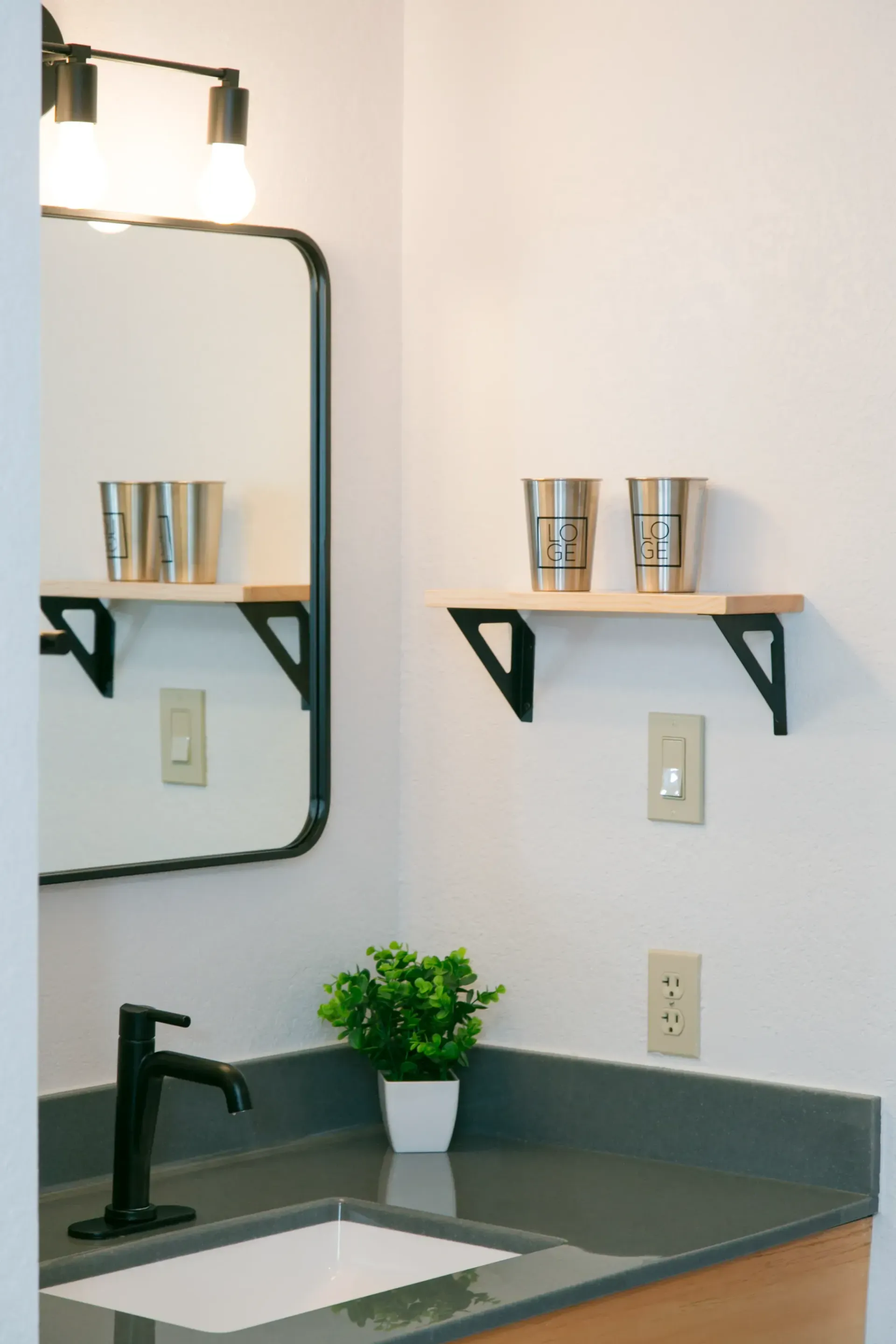 A modern bathroom sink with a black faucet, rectangular mirror, light wood wall shelves with metal cups, and a small plant.
