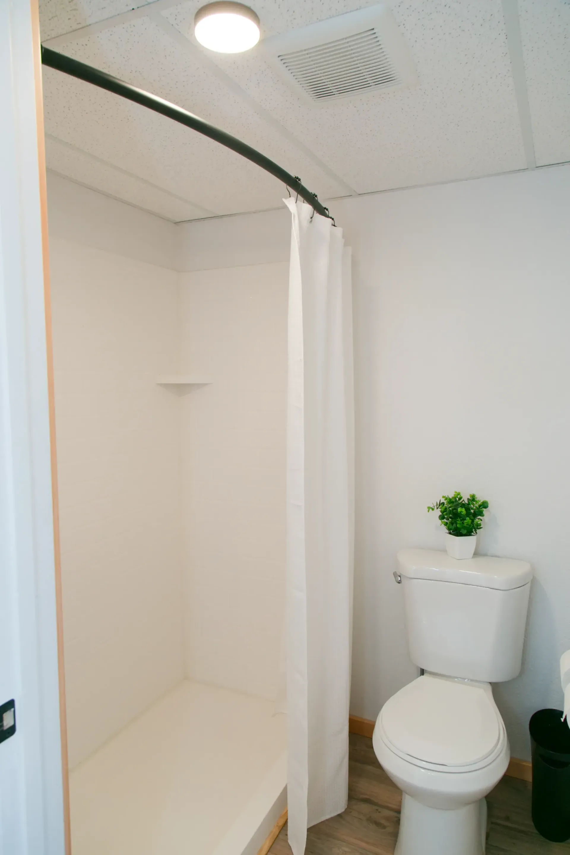 A white bathroom featuring a walk-in shower with a curved curtain rod and a white toilet with a small plant on the tank.