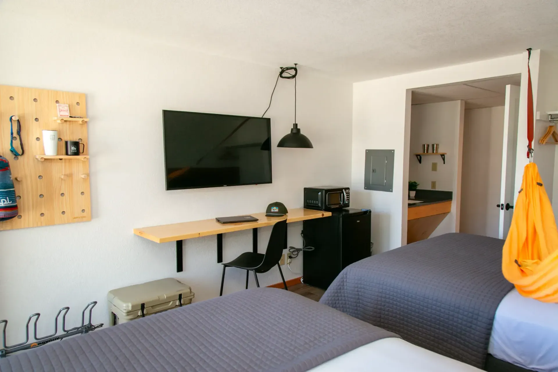 A hotel room with two beds, a mounted TV above a desk, a small refrigerator, and a bright yellow hammock chair.