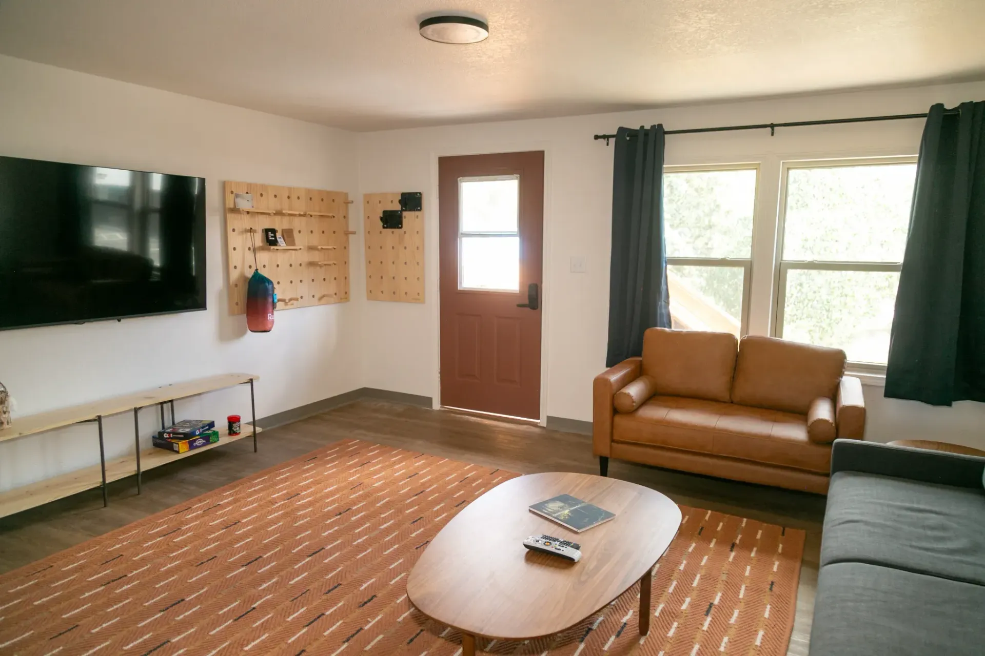 A living room with a large TV, tan leather loveseat, patterned rug, wooden coffee table, and a brown door.