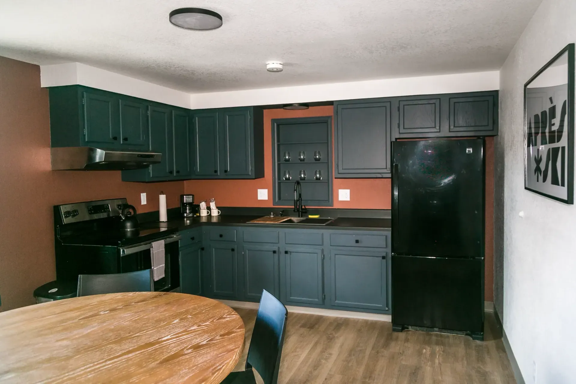A kitchen featuring dark blue cabinets, black appliances, dark countertops, and a wooden table in the foreground.
