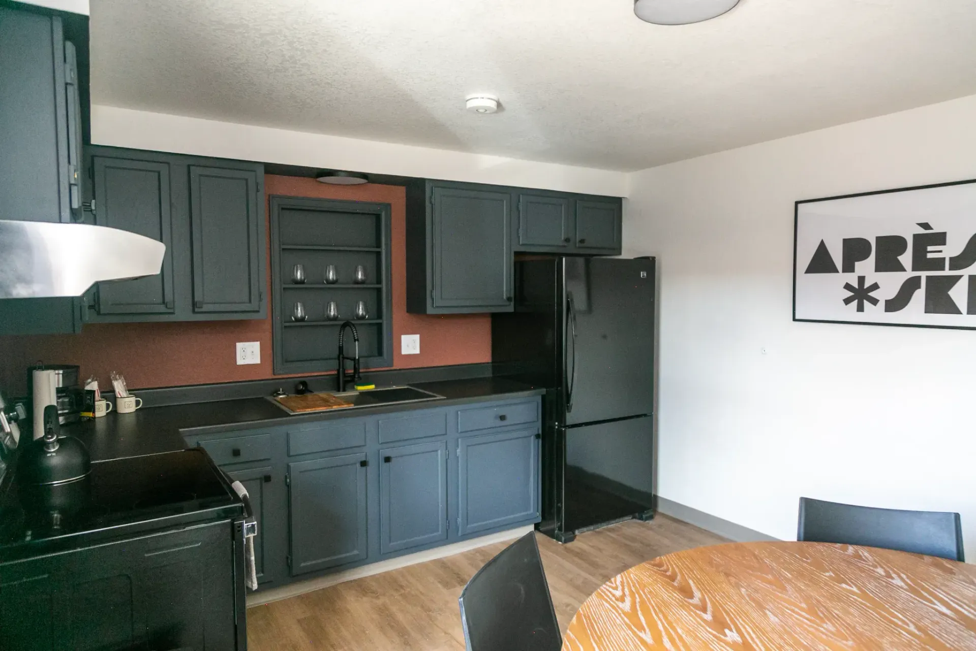 A kitchen featuring dark blue cabinets, black appliances, a wood-toned wall accent, and a round wooden dining table.