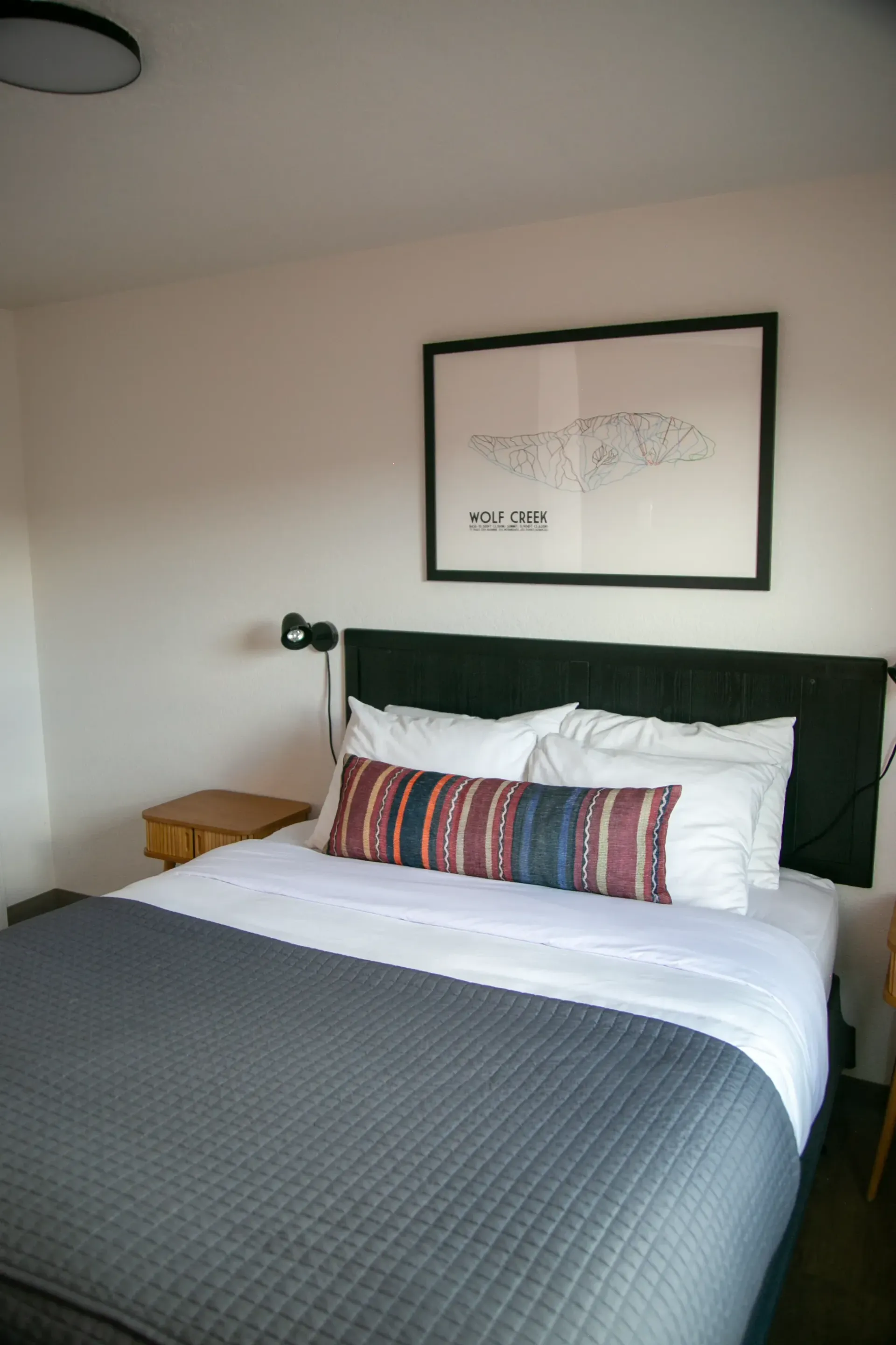 A modern bedroom features a bed with white sheets, a gray quilted coverlet, and a patterned lumbar pillow.