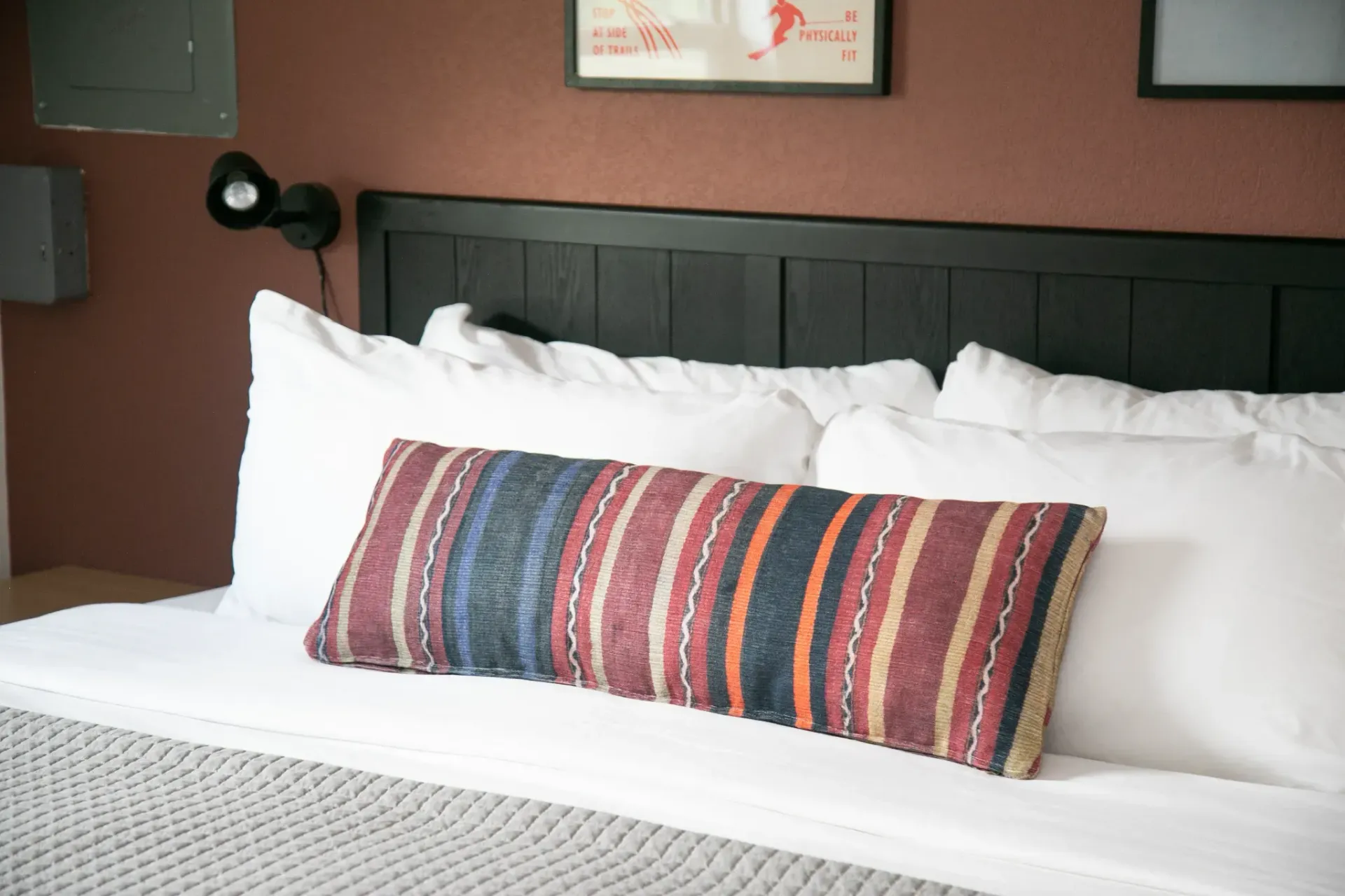 A close-up of a bed with white pillows, a long striped accent pillow, and a gray quilted blanket against a brown wall.